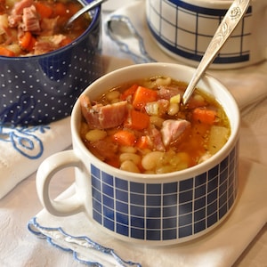 May include: Three bowls of white bean soup with ham and vegetables. The soup is in blue and white patterned bowls. Two bowls are on a white tablecloth with salt shakers and bread in the background.