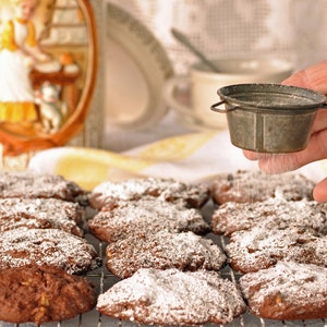 Puede incluir: Primer plano de galletas con chispas de chocolate espolvoreadas con azúcar glas. Las galletas están en una rejilla de alambre y una mano está espolvoreando azúcar glas desde un colador de metal.
