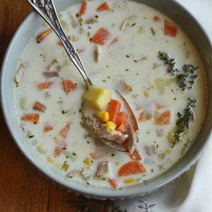 May include: A bowl of creamy white soup with chunks of potato, carrot, corn, and ham. The soup is garnished with fresh thyme sprigs. A silver spoon is resting in the bowl, filled with soup and vegetables.