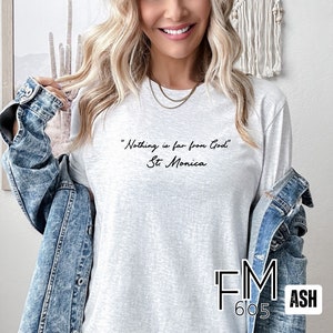 Puede incluir: Una camiseta de manga larga gris jaspeada con la cita "Nada est&aacute; lejos de Dios" - Santa M&oacute;nica impresa en negro en la parte delantera. La camiseta la lleva puesta una persona con el pelo largo y rubio. La camiseta est&aacute; etiquetada con "FM 605 ASH" en la esquina inferior derecha.