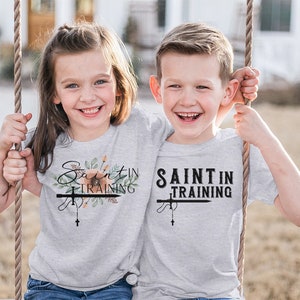 May include: Two children wearing light gray t-shirts with the text "Saint in Training". The shirts feature a cross and rosary design, with floral accents on one shirt. The children are smiling and holding onto a swing.