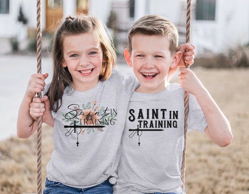 May include: Two children wearing light gray t-shirts with the text "Saint in Training". The shirts feature a cross and rosary design, with floral accents on one shirt. The children are smiling and holding onto a swing.