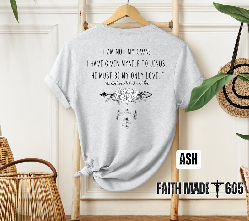 May include: A gray t-shirt with a quote from St. Kateri Tekakwitha: "I am not my own: I have given myself to Jesus. He must be my only love." The quote is printed in black text with a black and white arrow and flower design below it. The shirt has the words "ASH" and "FAITH MADE 605" printed on the bottom.