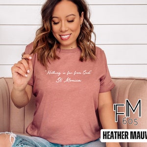 Puede incluir: Una camiseta de color malva con el texto "Nothing is far from God" y "St. Monica" en blanco. La camiseta est&aacute; hecha por FM 605.