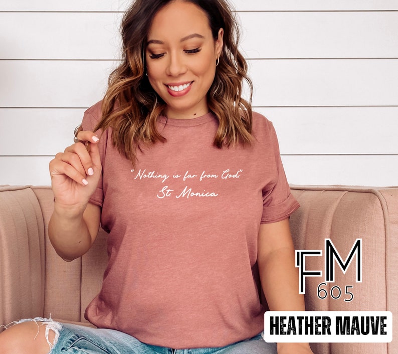 Puede incluir: Una camiseta de color malva con el texto "Nothing is far from God" y "St. Monica" en blanco. La camiseta est&aacute; hecha por FM 605.