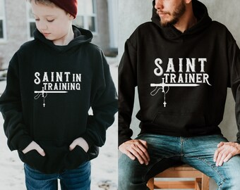 May include: Two black hoodies with white text. The smaller hoodie says "Saint in Training" and the larger hoodie says "Saint Trainer". Both hoodies feature a sword and rosary design.
