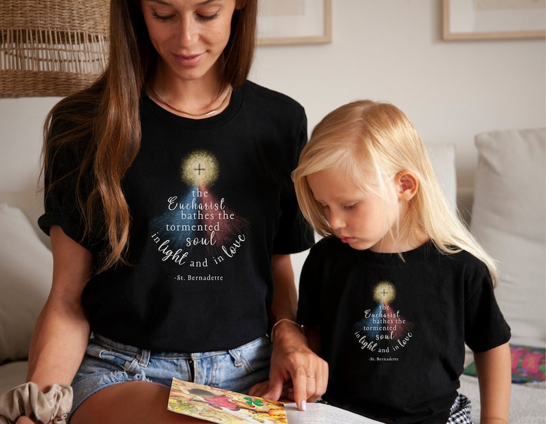 May include: Black t-shirts with a religious design. The shirts feature a graphic with a cross and the text "the Eucharist bathes the tormented soul in light and in love - St. Bernadette."