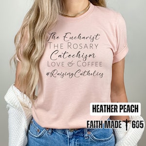 May include: Heather peach-colored t-shirt with the phrases "The Eucharist, The Rosary, Catechism, Love & Coffee" and the hashtag "#Raising Catholics" in black script. The shirt also has the text "Heather Peach" and "Faith Made 605".