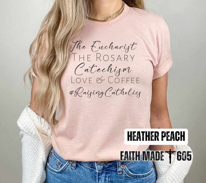 May include: Heather peach-colored t-shirt with the phrases "The Eucharist, The Rosary, Catechism, Love & Coffee" and the hashtag "#Raising Catholics" in black script. The shirt also has the text "Heather Peach" and "Faith Made 605".