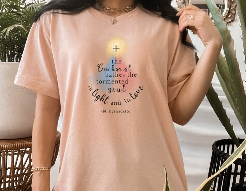 May include: A pale peach t-shirt featuring the quote "the Eucharist bathes the tormented soul in light and in love - St. Bernadette". The design includes a cross and colourful accents.