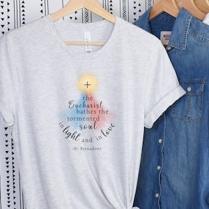 May include: A light grey t-shirt with the text "the Eucharist bathes the tormented soul in light and in love - St. Bernadette." The shirt is hanging on a wooden hanger with other shirts.