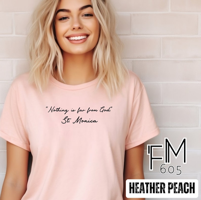 Puede incluir: Una camiseta rosa claro con la cita "Nada est&aacute; lejos de Dios" y el texto "Santa M&oacute;nica" impreso en negro. La camiseta est&aacute; etiquetada como "FM 605 Heather Peach".