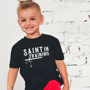 May include: A young child wearing a black t-shirt with the words "SAINT IN TRAINING" in white. The shirt features a graphic of a sword and rosary. The child is smiling and wearing red shorts.