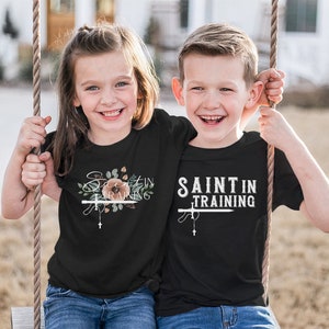 May include: Two children wearing black t-shirts with the text "Saint in Training". One shirt features floral accents and a cross, while the other has a simple design with a cross. Both children are smiling and holding onto a swing.