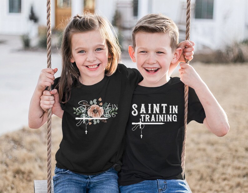 May include: Two children wearing black t-shirts with the text "Saint in Training". One shirt features floral accents and a cross, while the other has a simple design with a cross. Both children are smiling and holding onto a swing.