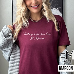Puede incluir: Una camiseta color granate con la cita "Nothing is far from God, St. Monica" impresa en blanco en la parte delantera. La camiseta est&aacute; siendo modelada por una persona con cabello rubio. El texto "FM 605 MAROON" es visible en la esquina inferior derecha de la imagen.
