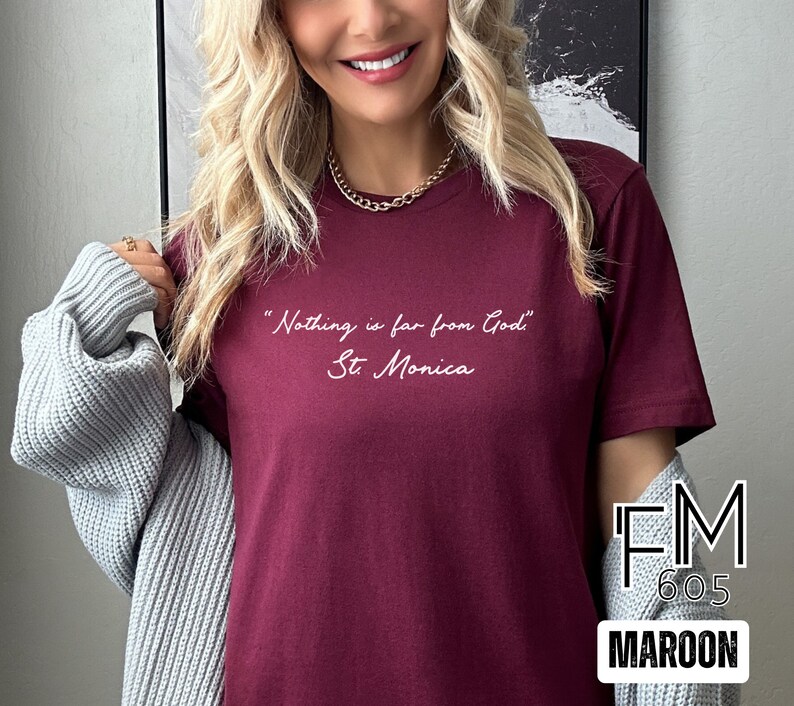 Puede incluir: Una camiseta color granate con la cita "Nothing is far from God, St. Monica" impresa en blanco en la parte delantera. La camiseta est&aacute; siendo modelada por una persona con cabello rubio. El texto "FM 605 MAROON" es visible en la esquina inferior derecha de la imagen.