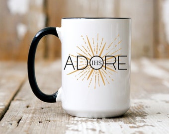 May include: White ceramic mug with a black handle and a gold design that says "ADORE IHS" with a sunburst pattern.