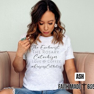 May include: A light gray t-shirt with black text that reads "The Eucharist, The Rosary, Catechism, Love & Coffee, #RaisingCatholics." The shirt also has the text "Faith Made 605" in a white box. The model is wearing the shirt.