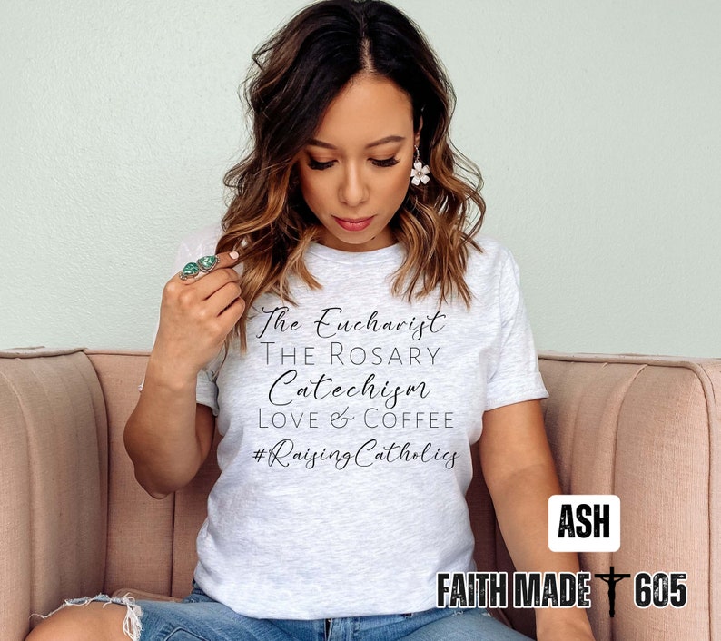 May include: A light gray t-shirt with black text that reads "The Eucharist, The Rosary, Catechism, Love & Coffee, #RaisingCatholics." The shirt also has the text "Faith Made 605" in a white box. The model is wearing the shirt.