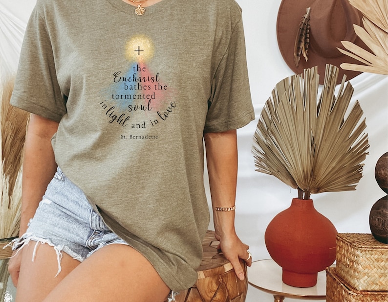 May include: An olive green t-shirt with a graphic design. The design includes the text "the Eucharist bathes the tormented soul in light and in love - St. Bernadette." The graphic features a cross and a rainbow-colored shape. The shirt is short-sleeved.