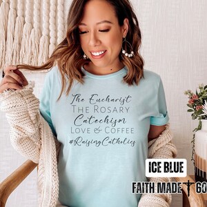 May include: Ice blue t-shirt with the text "The Eucharist, The Rosary, Catechism, Love & Coffee, #Raising Catholics." The shirt also has the text "ICE BLUE" and "FAITH MADE 605".