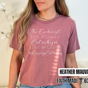 May include: Heather mauve t-shirt with white script text: "The Eucharist, The Rosary, Catechism, Love & Coffee, #Raising Catholics." The bottom right corner has the text "Heather Mauve" and "Faith Made 605" in a white rectangle.