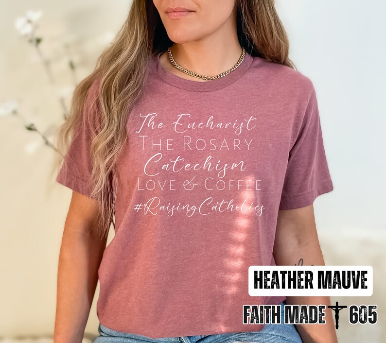 May include: Heather mauve t-shirt with white script text: "The Eucharist, The Rosary, Catechism, Love & Coffee, #Raising Catholics." The bottom right corner has the text "Heather Mauve" and "Faith Made 605" in a white rectangle.