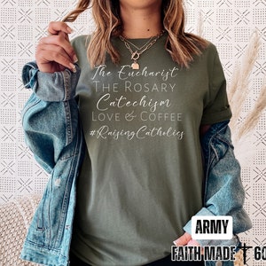 May include: Army green t-shirt with white script text: "The Eucharist, The Rosary, Catechism, Love & Coffee, #Raising Catholics." The shirt is paired with a denim jacket. The bottom right corner of the shirt has the text "ARMY" and "FAITH MADE 605."