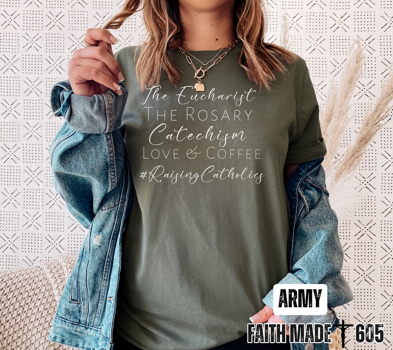 May include: Army green t-shirt with white script text: "The Eucharist, The Rosary, Catechism, Love & Coffee, #Raising Catholics." The shirt is paired with a denim jacket. The bottom right corner of the shirt has the text "ARMY" and "FAITH MADE 605."