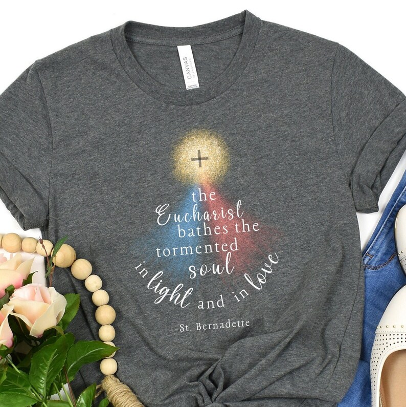 May include: Dark grey t-shirt featuring the quote "the Eucharist bathes the tormented soul in light and in love" and a cross design. The quote is attributed to St. Bernadette. The shirt has a crew neck.
