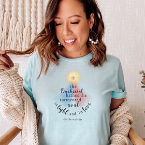 May include: Pale blue t-shirt featuring the quote "The Eucharist bathes the tormented soul in light and in love - St. Bernadette." The design includes a cross and watercolour accents.