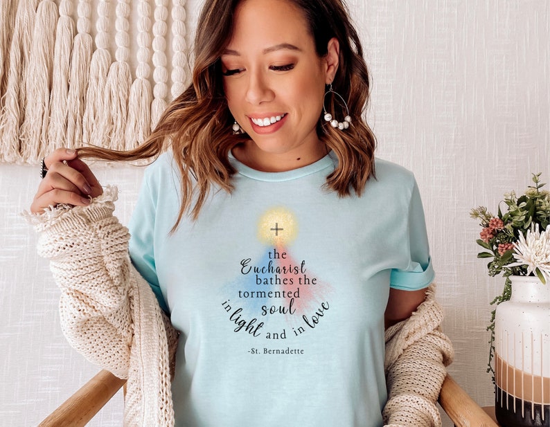 May include: Pale blue t-shirt featuring the quote "The Eucharist bathes the tormented soul in light and in love - St. Bernadette." The design includes a cross and watercolour accents.