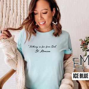 Puede incluir: Una camiseta azul claro con la cita "Nada est&aacute; lejos de Dios" y "Santa M&oacute;nica" escrita en cursiva negra. La camiseta est&aacute; en una modelo y el fondo es una pared blanca.