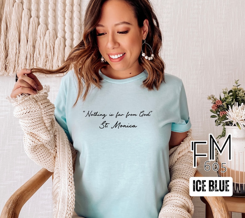Puede incluir: Una camiseta azul claro con la cita "Nada est&aacute; lejos de Dios" y "Santa M&oacute;nica" escrita en cursiva negra. La camiseta est&aacute; en una modelo y el fondo es una pared blanca.