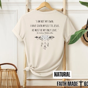 May include: A beige t-shirt with a quote about faith and love. The quote reads "I am not my own: I have given myself to Jesus. He must be my only love."  The quote is attributed to St. Kateri Tekakwitha. The shirt also features a floral design with an arrow and feathers.