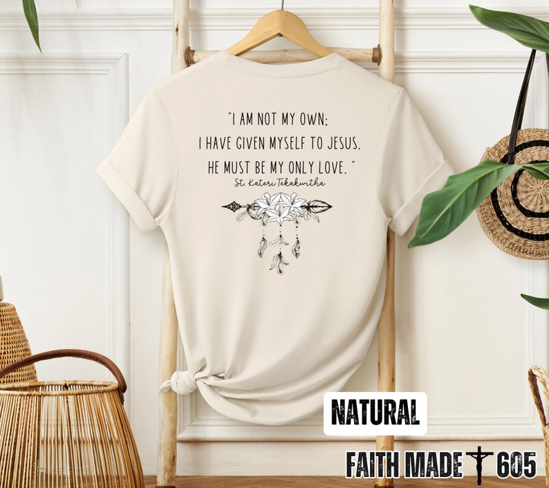 May include: A beige t-shirt with a quote about faith and love. The quote reads "I am not my own: I have given myself to Jesus. He must be my only love."  The quote is attributed to St. Kateri Tekakwitha. The shirt also features a floral design with an arrow and feathers.
