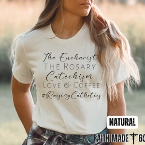 May include: Cream-colored t-shirt with the phrases "The Eucharist, The Rosary, Catechism, Love & Coffee, #RaisingCatholics" in black script. The shirt also has the word "NATURAL" and the text "FAITH MADE 605" in a white rectangle.