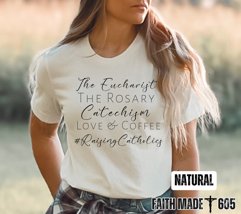 May include: Cream-colored t-shirt with the phrases "The Eucharist, The Rosary, Catechism, Love & Coffee, #RaisingCatholics" in black script. The shirt also has the word "NATURAL" and the text "FAITH MADE 605" in a white rectangle.
