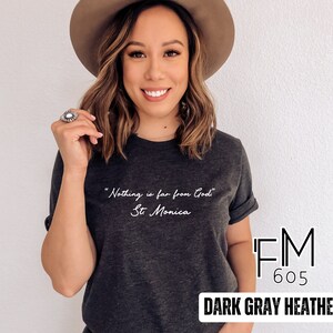Puede incluir: Camiseta de color gris oscuro jaspeado con la cita "Nada est&aacute; lejos de Dios" - Santa M&oacute;nica. La camiseta tiene una impresi&oacute;n blanca y el logotipo de la marca "FM 605" es visible en la esquina inferior derecha.
