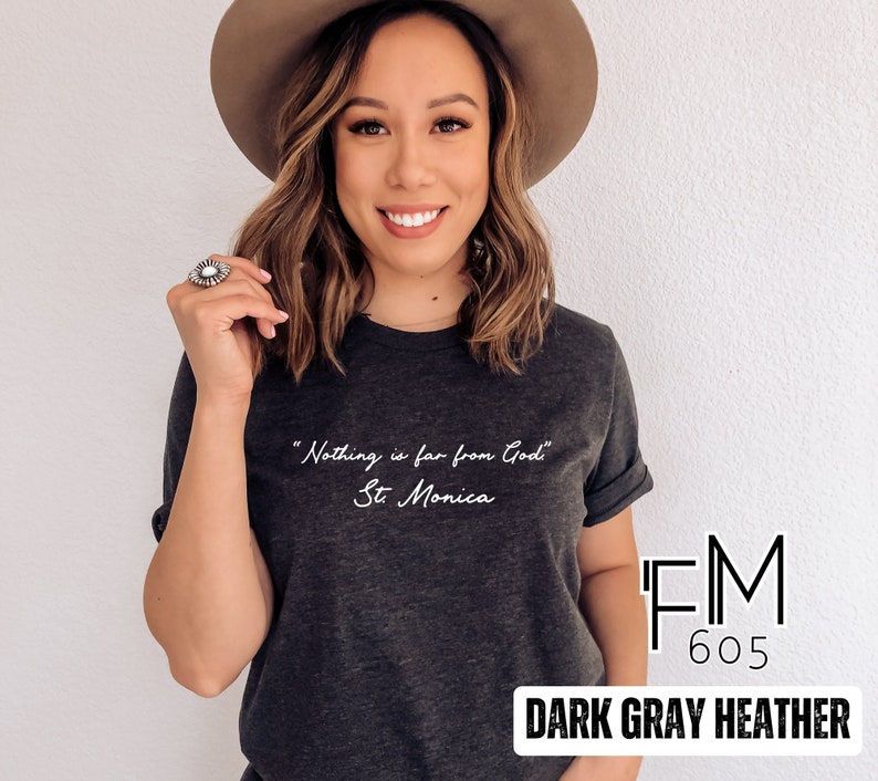 Puede incluir: Camiseta de color gris oscuro jaspeado con la cita "Nada est&aacute; lejos de Dios" - Santa M&oacute;nica. La camiseta tiene una impresi&oacute;n blanca y el logotipo de la marca "FM 605" es visible en la esquina inferior derecha.
