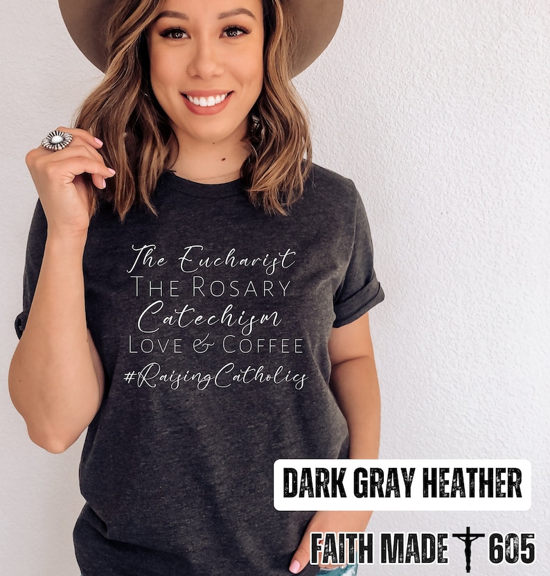 May include: Dark gray heather t-shirt with white text: "The Eucharist, The Rosary, Catechism, Love & Coffee, #Raising Catholics." The bottom right corner reads "FAITH MADE 605".