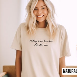 Puede incluir: Una camiseta beige con la cita "Nada est&aacute; lejos de Dios" - Santa M&oacute;nica impresa en negro en la parte delantera. La palabra "NATURAL" est&aacute; impresa en negro en la esquina inferior derecha de la camiseta.