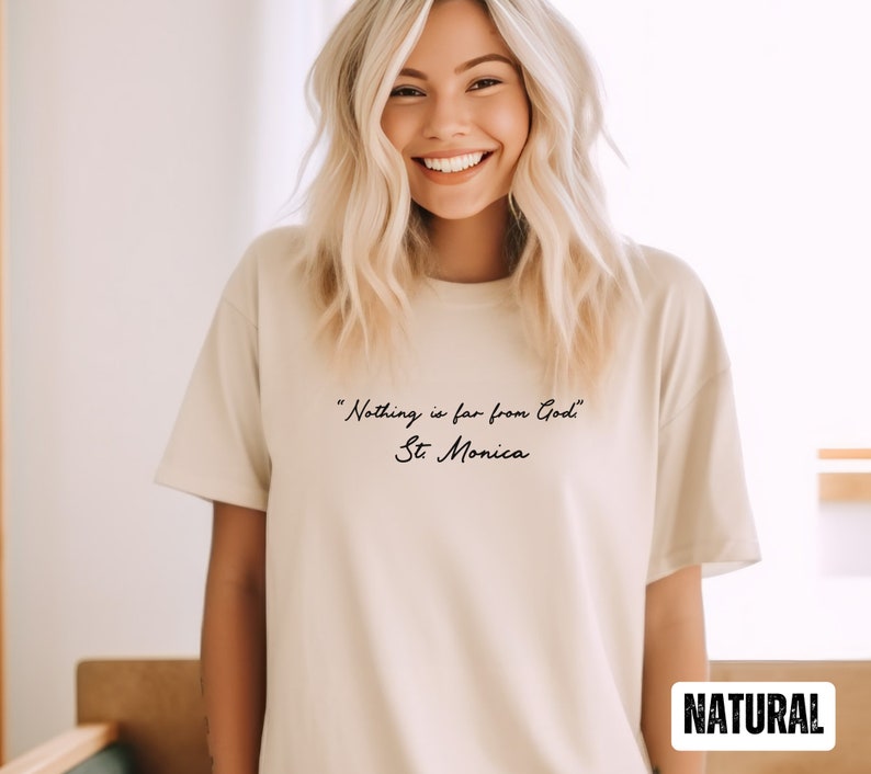 Puede incluir: Una camiseta beige con la cita "Nada est&aacute; lejos de Dios" - Santa M&oacute;nica impresa en negro en la parte delantera. La palabra "NATURAL" est&aacute; impresa en negro en la esquina inferior derecha de la camiseta.