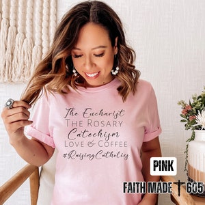 May include: A light pink t-shirt with the phrases "The Eucharist, The Rosary, Catechism, Love & Coffee" and the hashtag "#RaisingCatholics" in black script. The words "PINK" and "FAITH MADE 605" are also visible.