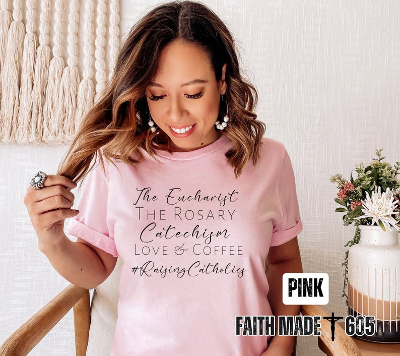 May include: A light pink t-shirt with the phrases "The Eucharist, The Rosary, Catechism, Love & Coffee" and the hashtag "#RaisingCatholics" in black script. The words "PINK" and "FAITH MADE 605" are also visible.