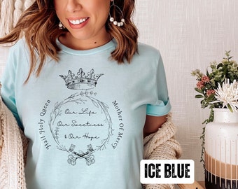 May include: Light blue t-shirt with a black graphic design featuring a crown, floral wreath, and the text "Hail Holy Queen, Our Life, Our Sweetness & Our Hope, Mother of Mercy". The text "ICE BLUE" and "FAITH MADE 605" are also visible.
