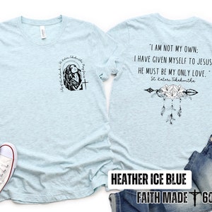 May include: A light blue t-shirt with a black graphic on the front and a black graphic with text on the back. The text on the back reads "I am not my own: I have given myself to Jesus. He must be my only love."  St. Kateri Tekakwitha. The t-shirt is displayed on a model with a pair of white sneakers and blue jeans.