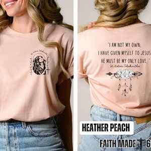 May include: A heather peach colored t-shirt with a graphic of a woman praying on the front and a quote from St. Kateri Tekakwitha on the back. The quote reads "I am not my own: I have given myself to Jesus. He must be my only love." The back of the shirt also features a graphic of an arrow with lilies and feathers. The shirt is labeled "Faith Made 605".