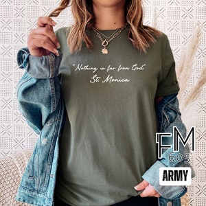 Puede incluir: Una camiseta verde con la cita "Nada est&aacute; lejos de Dios" y el nombre "Santa M&oacute;nica" escrito en blanco. La camiseta la lleva puesta una persona. La imagen tambi&eacute;n incluye una chaqueta vaquera y el texto "FM 605 ARMY".